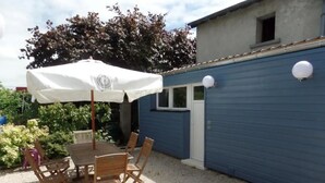 Eine Terrasse im Freien mit Tisch und Stühlen, einem weißen Sonnenschirm und einem blauen Gebäude im Hintergrund.