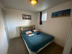 A single bed with a wooden headboard, a window with curtains, a painting of lighthouses, and a small shelf with a painting.
