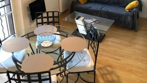 Un salon moderne avec une table ronde en verre, des chaises en métal noir, un canapé gris agrémenté de coussins jaunes, un luminaire suspendu et des œuvres d’art encadrées accrochées au mur.