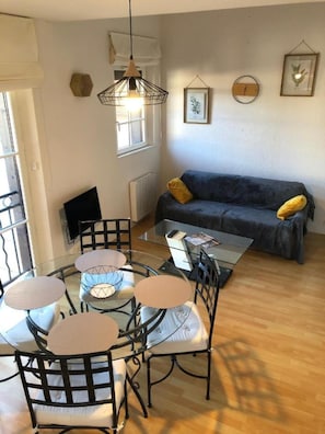 Un salon moderne avec une table ronde en verre, des chaises en métal noir, un canapé gris agrémenté de coussins jaunes, un luminaire suspendu et des œuvres d’art encadrées accrochées au mur.