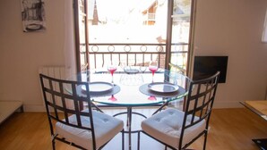 Une table ronde en verre, accompagnée de deux chaises, un balcon doté d’une rambarde en métal et un téléviseur fixé au mur.