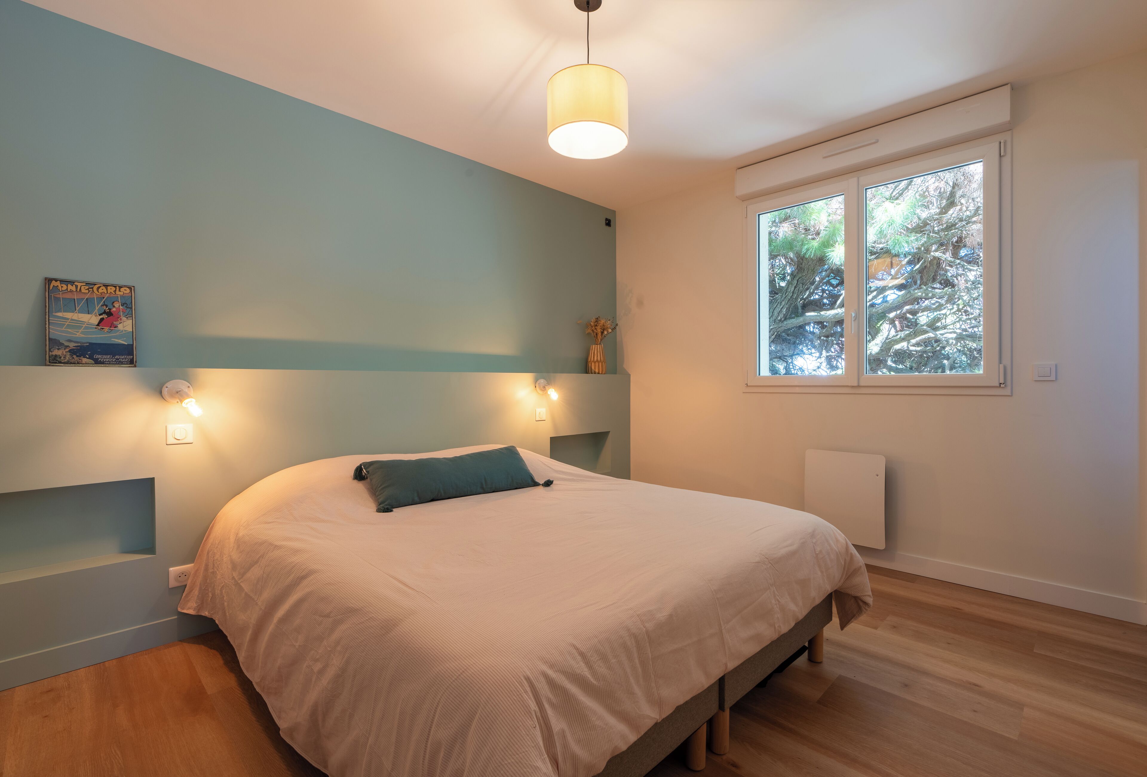 Une chambre à coucher avec un lit, une fenêtre donnant sur des arbres, une étagère fixée au mur et un luminaire fixé au mur.