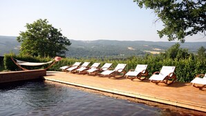 Terraza de madera con sillas de descanso y una hamaca junto a una piscina con vistas panorámicas.
