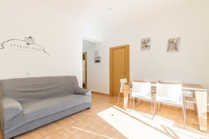 Un salon avec un canapé gris, une table à manger en bois avec des chaises, une porte en bois et un panneau indiquant la présence d’une connexion Wi-Fi, fixé au mur.