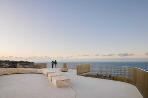 A waterfront terrace with a curved wooden deck, seating areas, and a railing overlooking the sea.