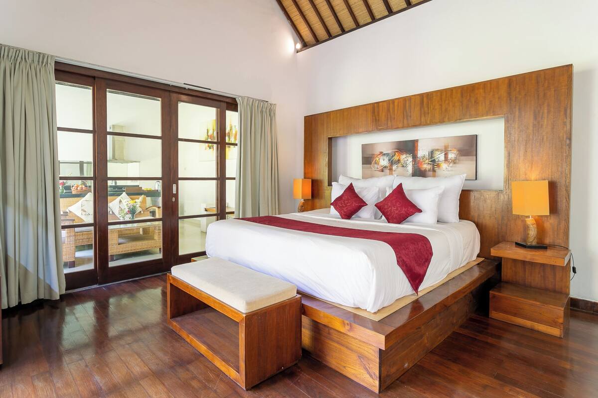 A spacious bedroom with a large bed, wooden headboard, and a sliding glass door leading to a balcony.