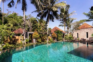 A clear swimming pool surrounded by lush greenery and traditional-style buildings.