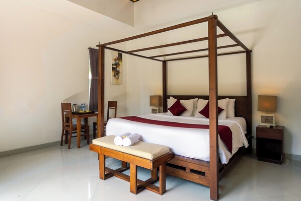 A four-poster bed with a wooden frame, a bench with towels, a bedside table with a lamp, and a small table with bottles and a kettle.