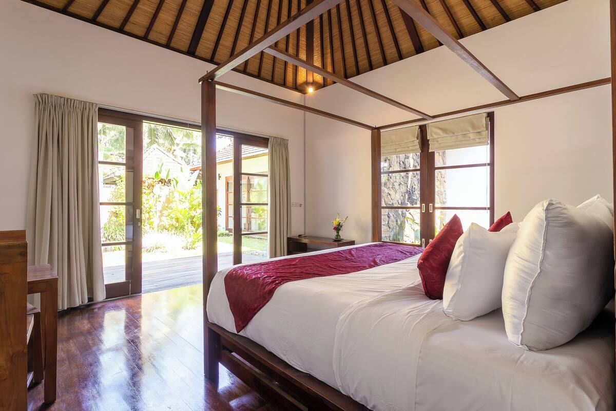 A bedroom with a wooden ceiling, a large bed with white and red pillows, a wooden nightstand, and a view of greenery outside.