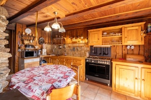 A rustic kitchen with wooden cabinets, a stove, a microwave, and a table with a quilted tablecloth. - Vacation Home “Chalet d'Alpage Authentique” with Wi-Fi (Queige)