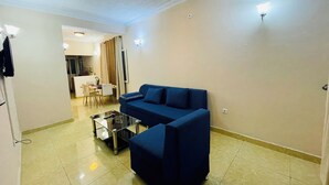 A living room with a blue sofa, a glass coffee table, and a dining area in the background. - Cosy Appartement Dans Quartier Calme à Proximité du Parc Aquatique (Kinshasa)
