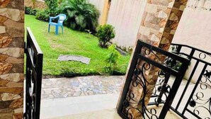 A view from a balcony overlooking a stone wall, a grassy area with a blue chair, and a tiled floor. - Cosy Appartement Dans Quartier Calme à Proximité du Parc Aquatique (Kinshasa)