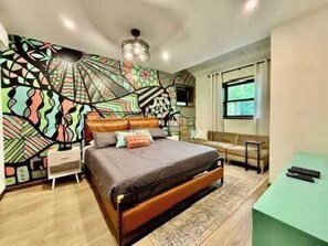 A bedroom with a bed, a nightstand, a desk, a chair, and a wall with a geometric pattern.