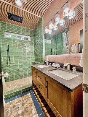 A bathroom with green tiled walls, a wooden vanity with two sinks, a mirror, and a shower area.