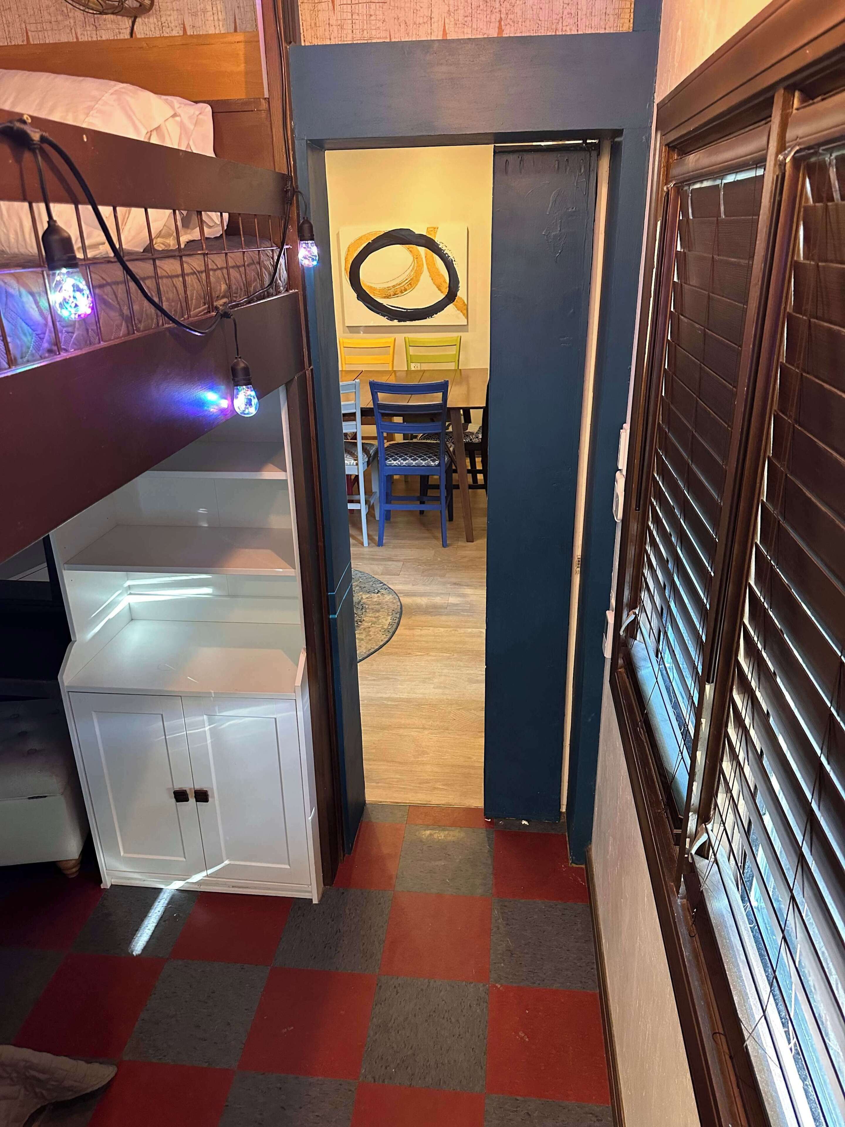A narrow hallway with a white cabinet, a dining table with chairs, and a window with blinds.