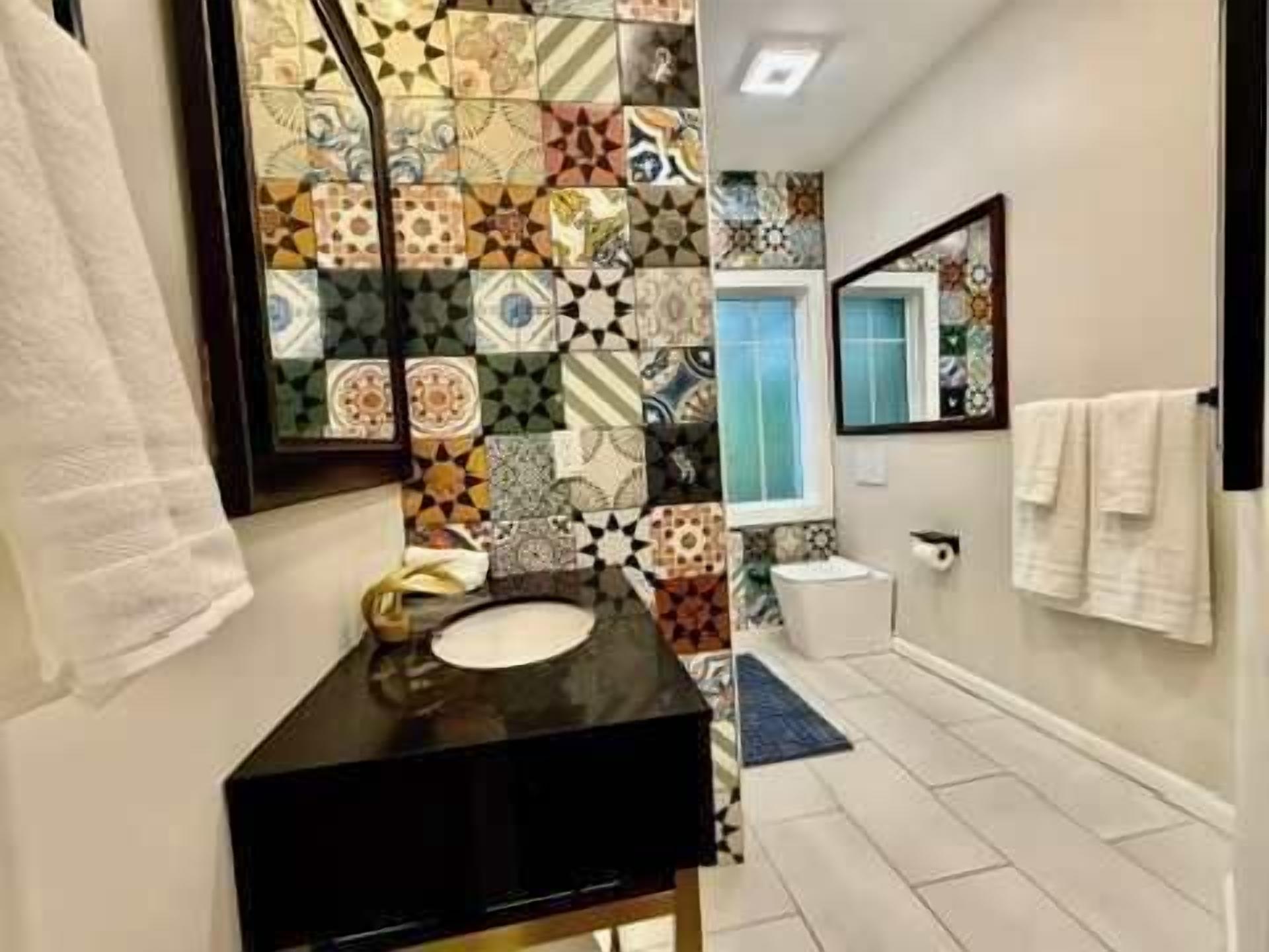 Une salle de bain avec un mur recouvert de carreaux décoratifs, un meuble-lavabo noir, un lavabo blanc et un miroir.