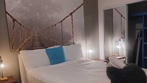 A bedroom with a large bed, a nightstand, a lamp, a mirror, and a wall-mounted artwork.