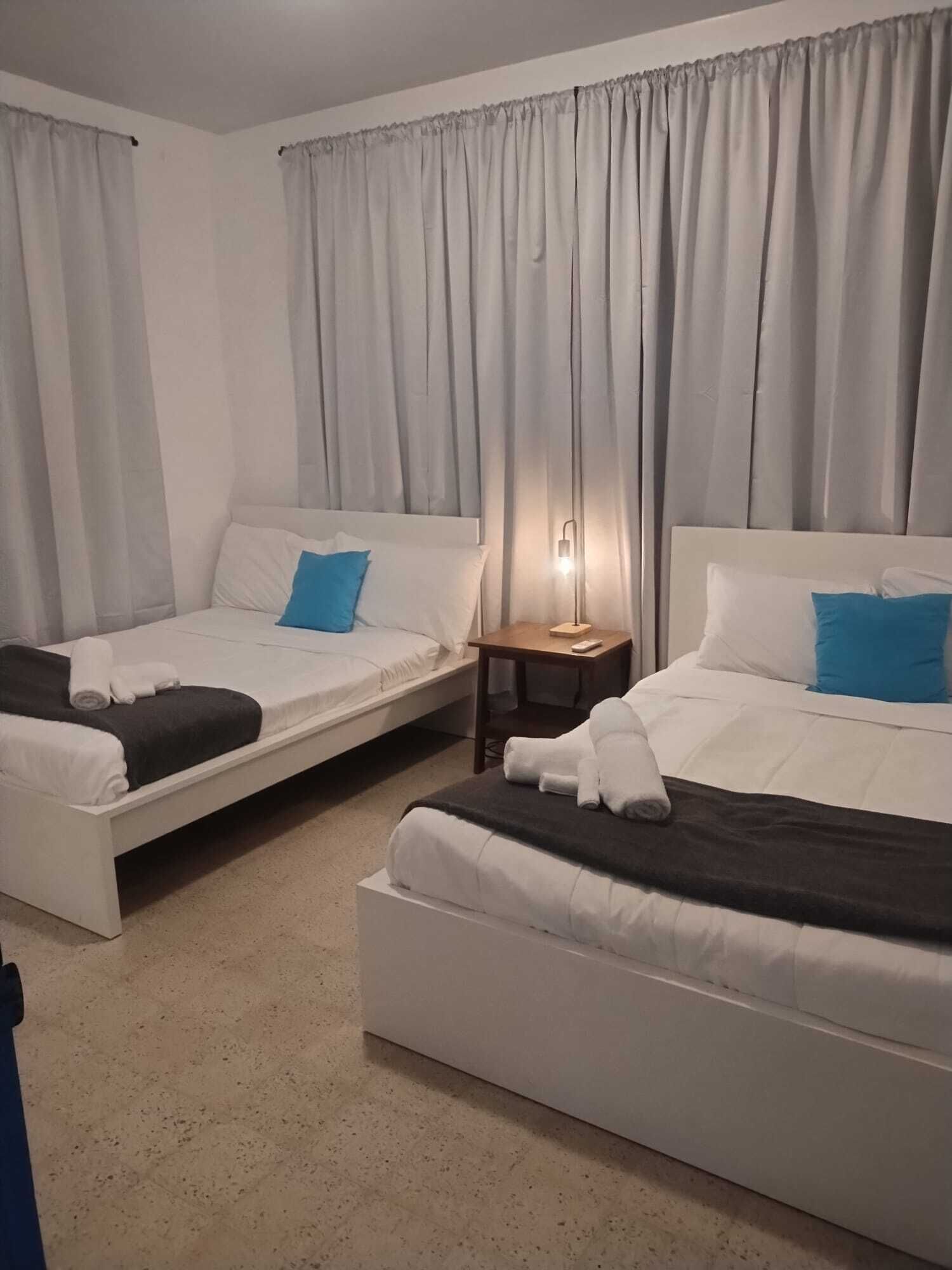 Two single beds with white bedding and blue pillows, a small wooden nightstand with a lamp, and a large window with grey curtains.