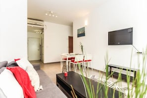 Ein modernes Wohnzimmer mit einem grauen Sofa, einem Esstisch mit roten Kissen, einem schwarzen Couchtisch und einem an der Wand befestigten Fernseher.
