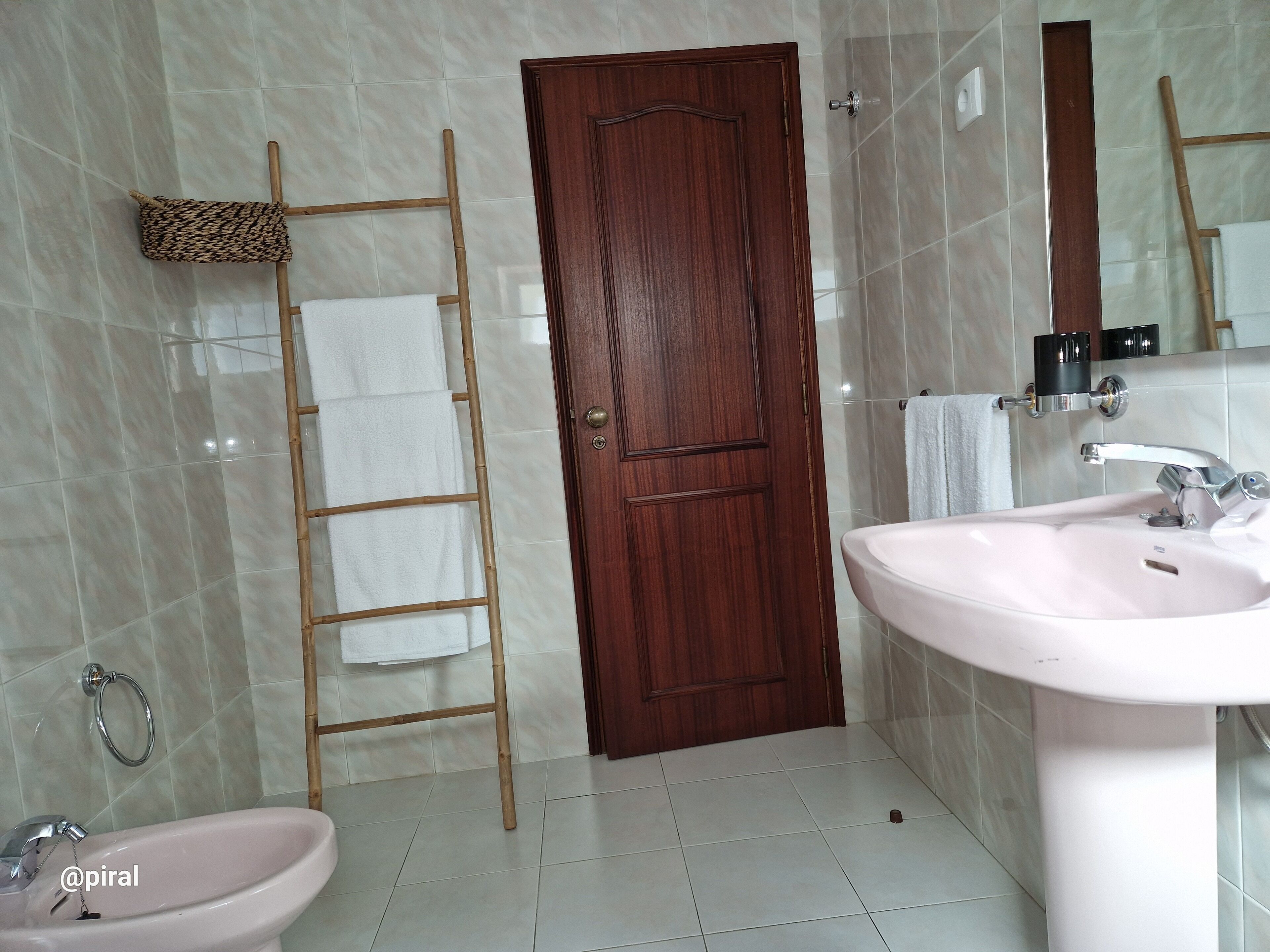 Un baño con puerta de madera, lavamanos blanco, espejo y perchero.