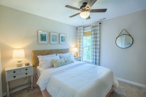 A neatly made bed with white bedding, a bedside lamp, and a nightstand with drawers. There is a ceiling fan, a window with curtains, and a wall-mounted mirror.