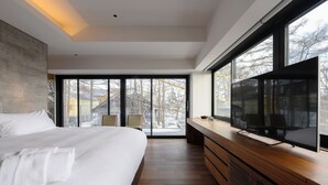 Ein modernes Schlafzimmer mit einem großen Bett, einem Fernseher auf einer Holzkonsole und einem Blick ins Freie durch eine Schiebetür.