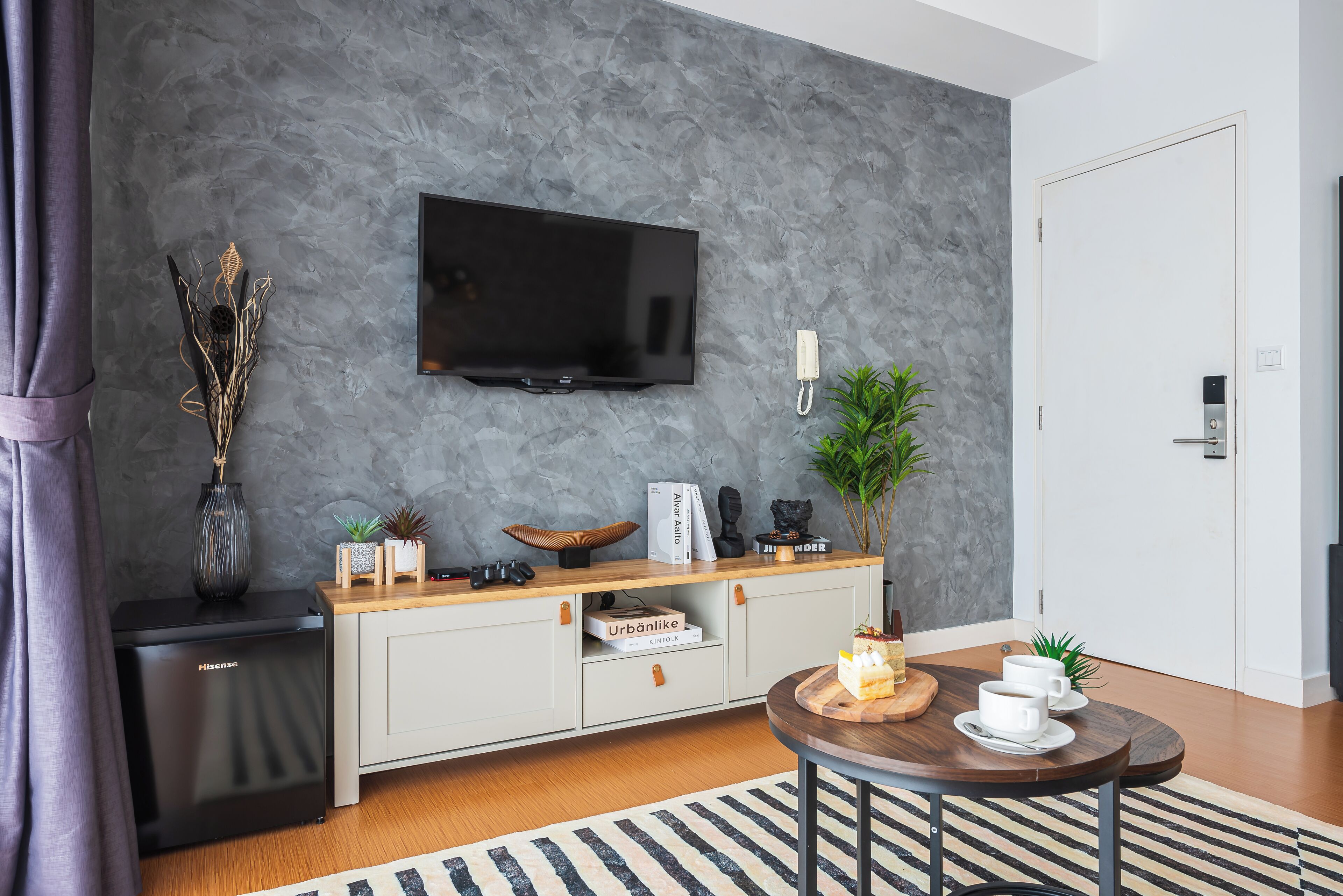 A modern living room with a grey textured wall, a mounted TV, a small fridge, a wooden console table with decorative items, and a round coffee table with cups and a cake.