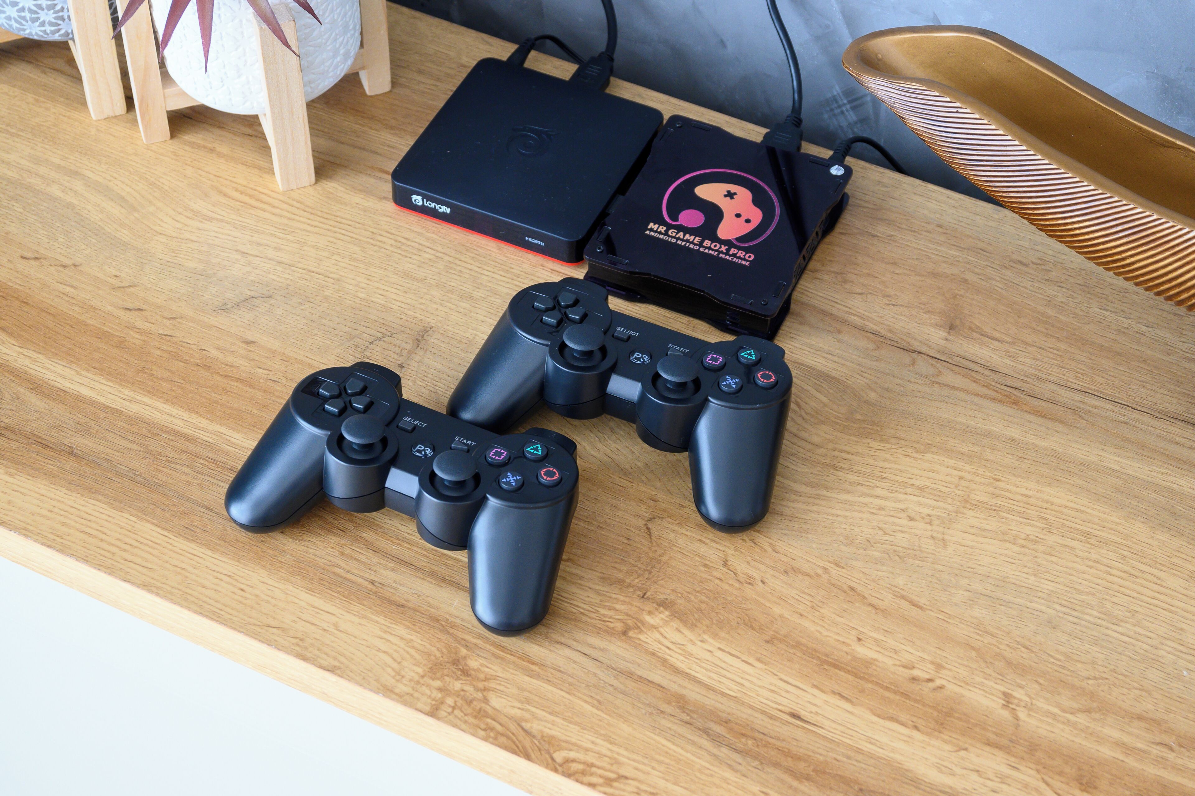 A wooden table with two black game controllers, a black gaming console, and a black box with a pink and black design.