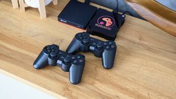 A wooden table with two black game controllers, a black gaming console, and a black box with a pink and black design.