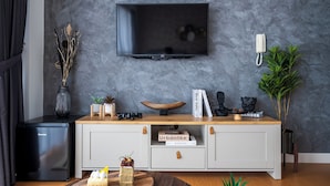 Una sala moderna con un televisor de pantalla plana montado en una pared gris con textura, un refrigerador mini negro, una mesa consola de madera con objetos decorativos y una planta en maceta.