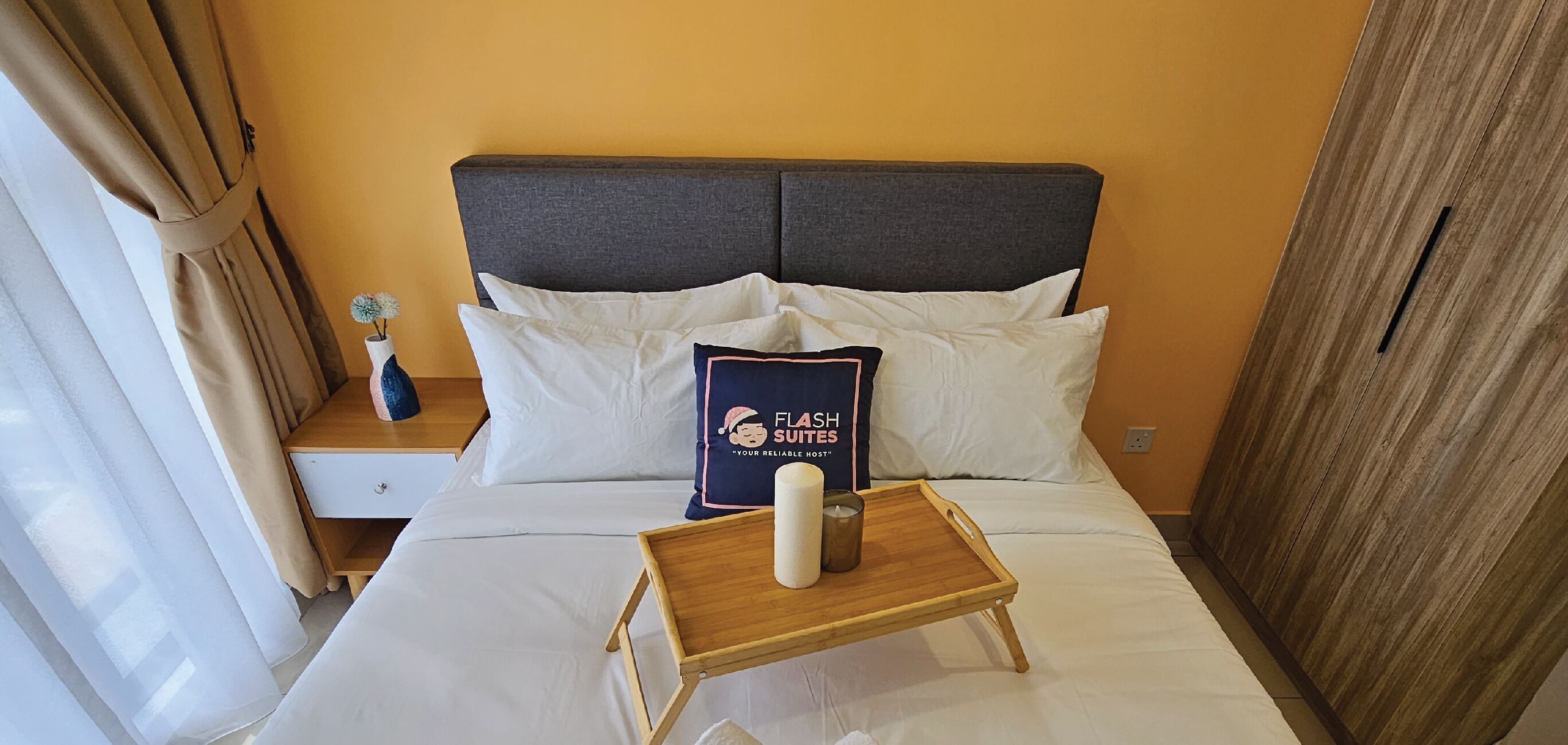 Una habitación de hotel con una cama, una mesita de noche, una mesilla auxiliar, una bandeja de madera con una vela y un cojín con un logotipo.