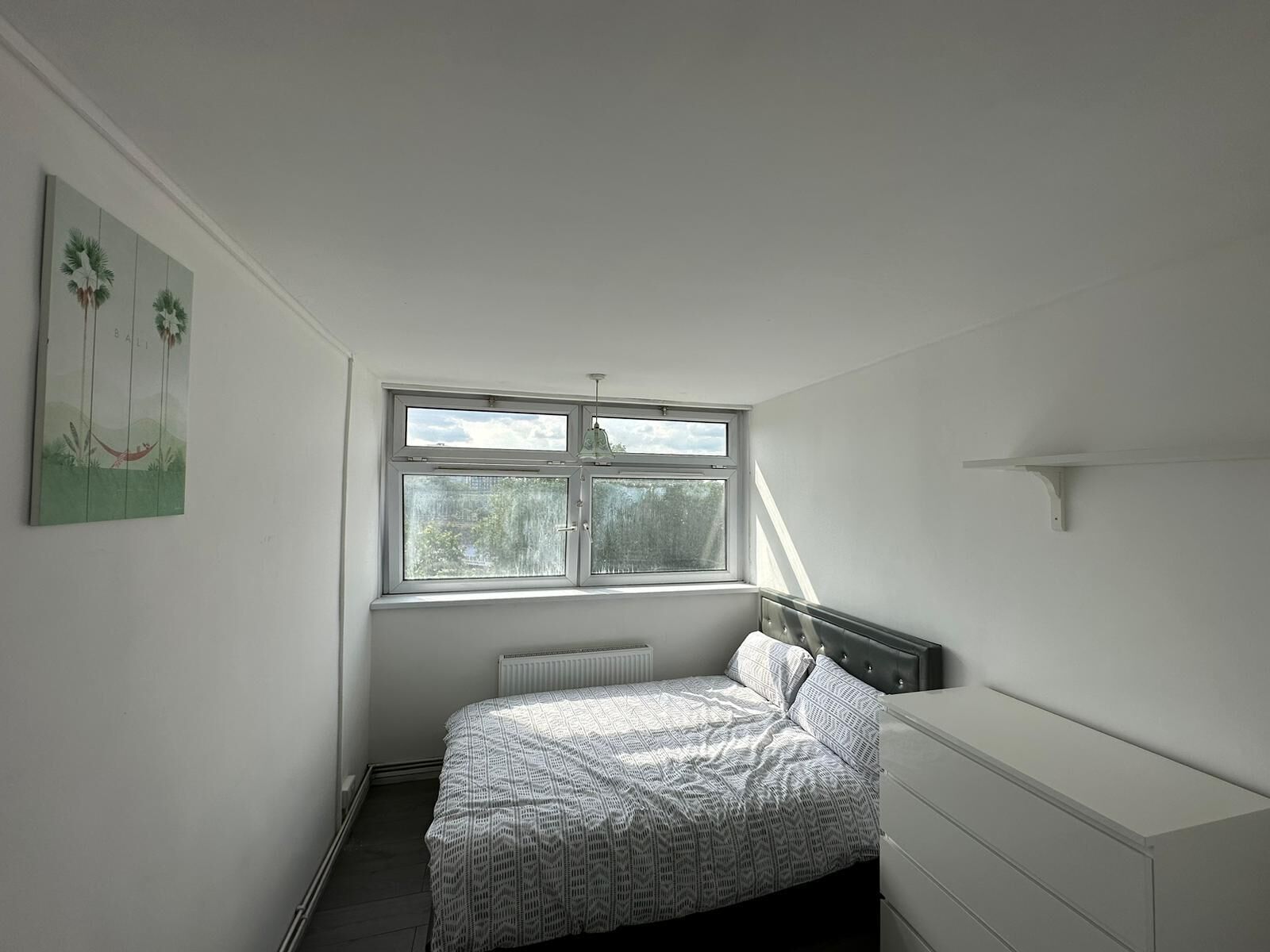 Une chambre à coucher avec un lit, un radiateur, une fenêtre et une étagère fixée au mur.