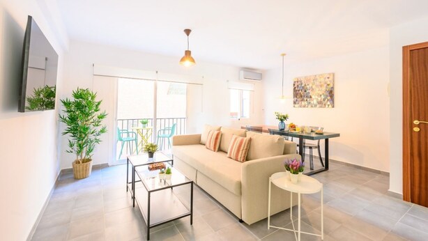 A modern living room with a sofa, coffee table, and dining table. There is a large window with a view of the outside, a ceiling fan, and a painting on the wall. - 2 bedroom, city and sea (Fuengirola)