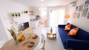 Un salon moderne avec un canapé bleu, une table à manger où est servi le petit-déjeuner, une cuisine en arrière-plan et un balcon avec vue.