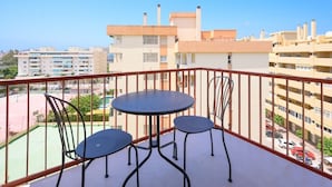 Un balcon avec une table ronde et deux chaises, donnant sur un quartier résidentiel avec des immeubles et des voitures garées.
