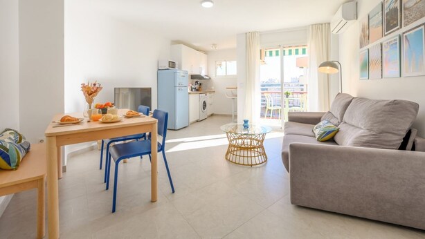 A modern living room with a sofa, a dining table with chairs, a kitchen area, and a view of the city through the window. - Terrace, swimming pool, parking (Fuengirola)