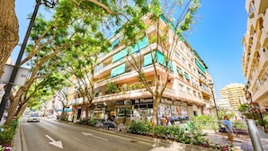 Un edificio de varios pisos con balcones, una calle al frente y una acera con árboles.