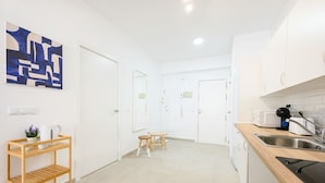 Una cocina moderna con armarios blancos, encimera de madera y un lavamanos. Se observa un taburete pequeño y una estantería con una tetera y una planta.