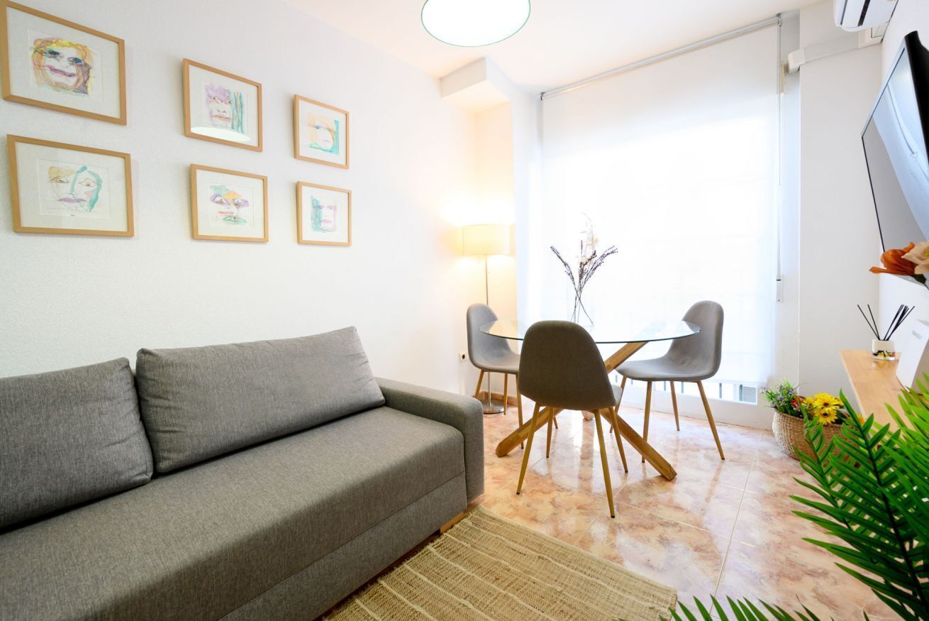 A living room with a grey sofa, a dining table with two chairs, and framed artwork on the wall.