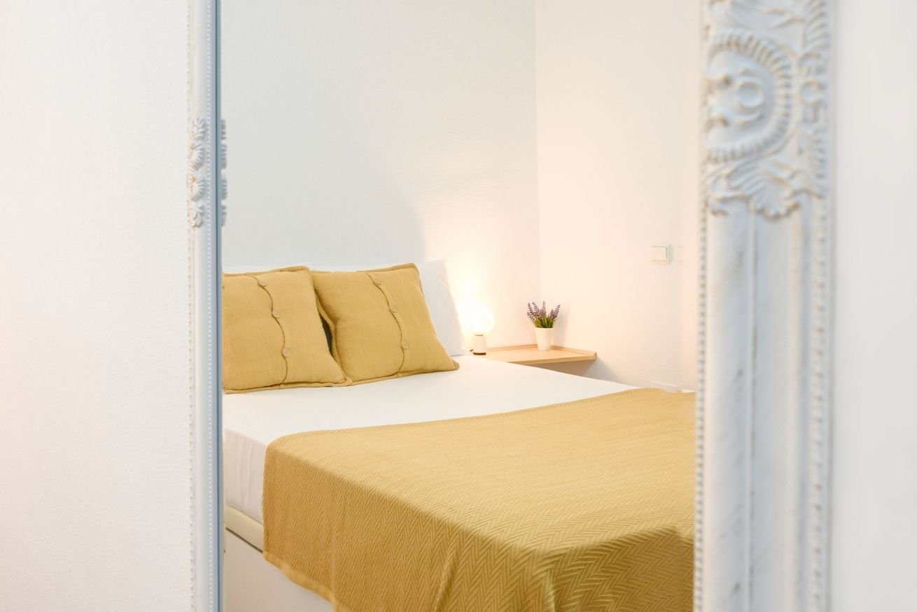 A neatly made bed with a yellow bedspread and pillows, a bedside table with a lamp and a small plant, and a decorative mirror with an ornate frame.