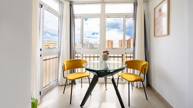 Habitación de esquina con una mesa de vidrio y sillas amarillas, una planta en maceta y vistas de edificios a través de ventanas amplias.
