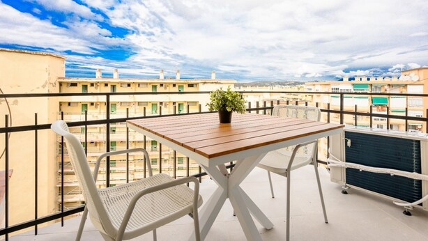 A balcony with a wooden table and two white chairs, a potted plant, and a view of buildings under a partly cloudy sky. - New Studio. Terrace & pool by 10ToSea (Fuengirola)