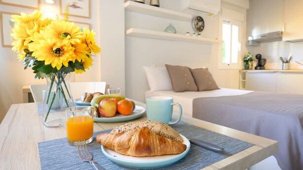 En frukostuppsättning med en croissant, apelsinjuice och en tallrik med frukt på ett bord, med en säng och ett kök i bakgrunden.