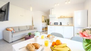 Une cuisine moderne avec des meubles blancs, une table à manger où sont disposés des aliments pour le petit-déjeuner, et un coin salon avec un canapé et un téléviseur.