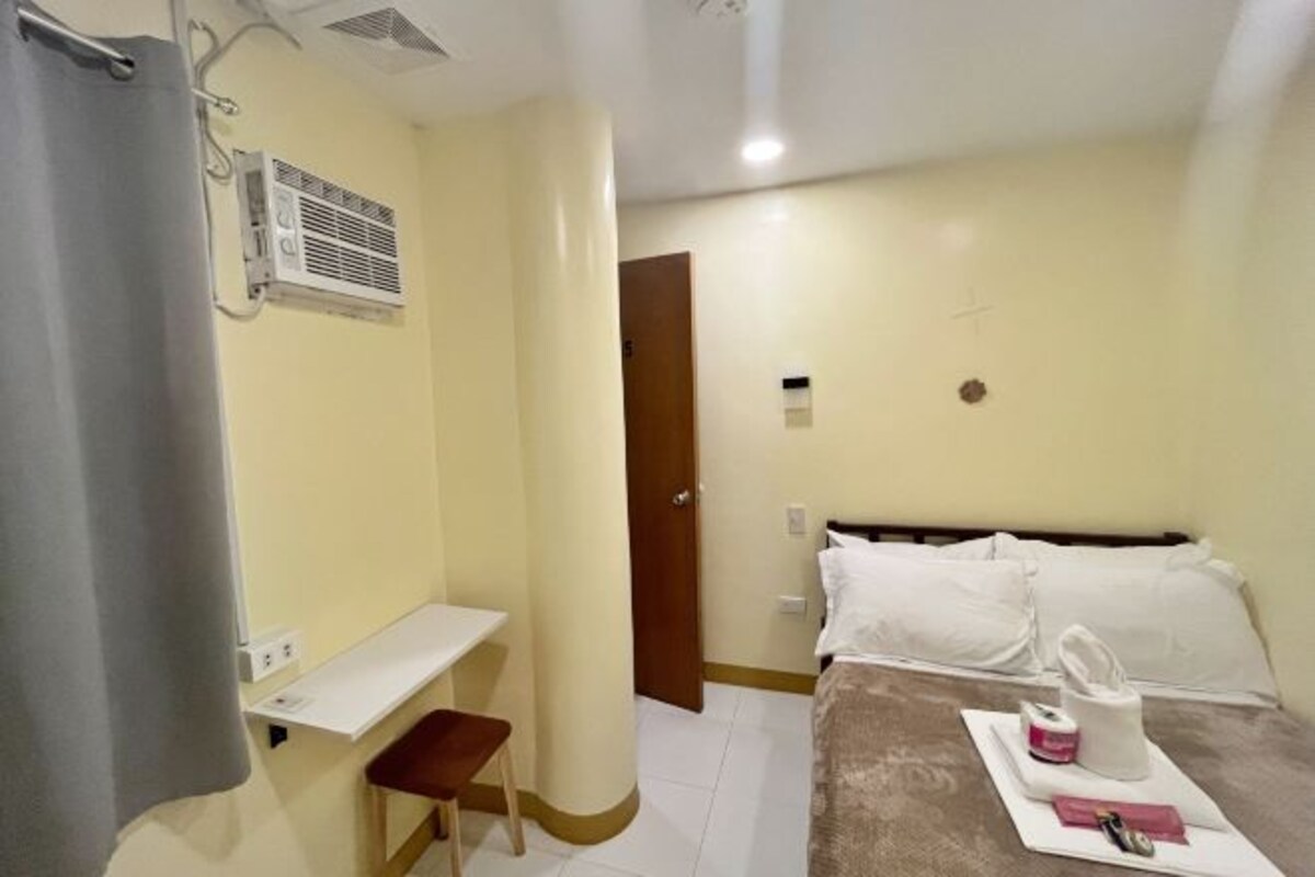 A hotel room with a bed, a desk, a chair, a curtain, an air conditioning unit, and a door.