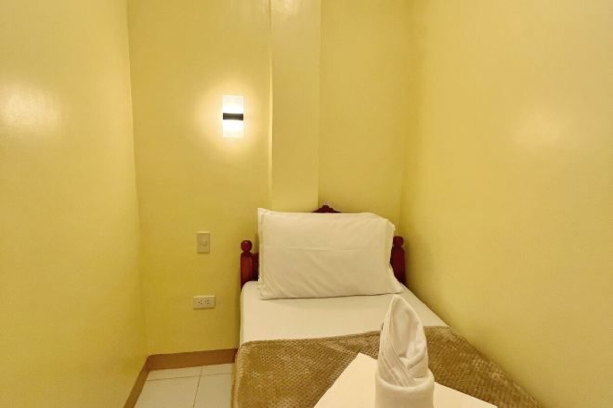 A small, yellow-walled room with a single bed, a nightstand, and a wall-mounted light.
