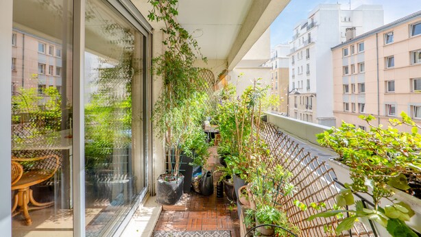 Un balcon agrémenté de plantes en pot, une chaise en osier et une vue sur les immeubles voisins.