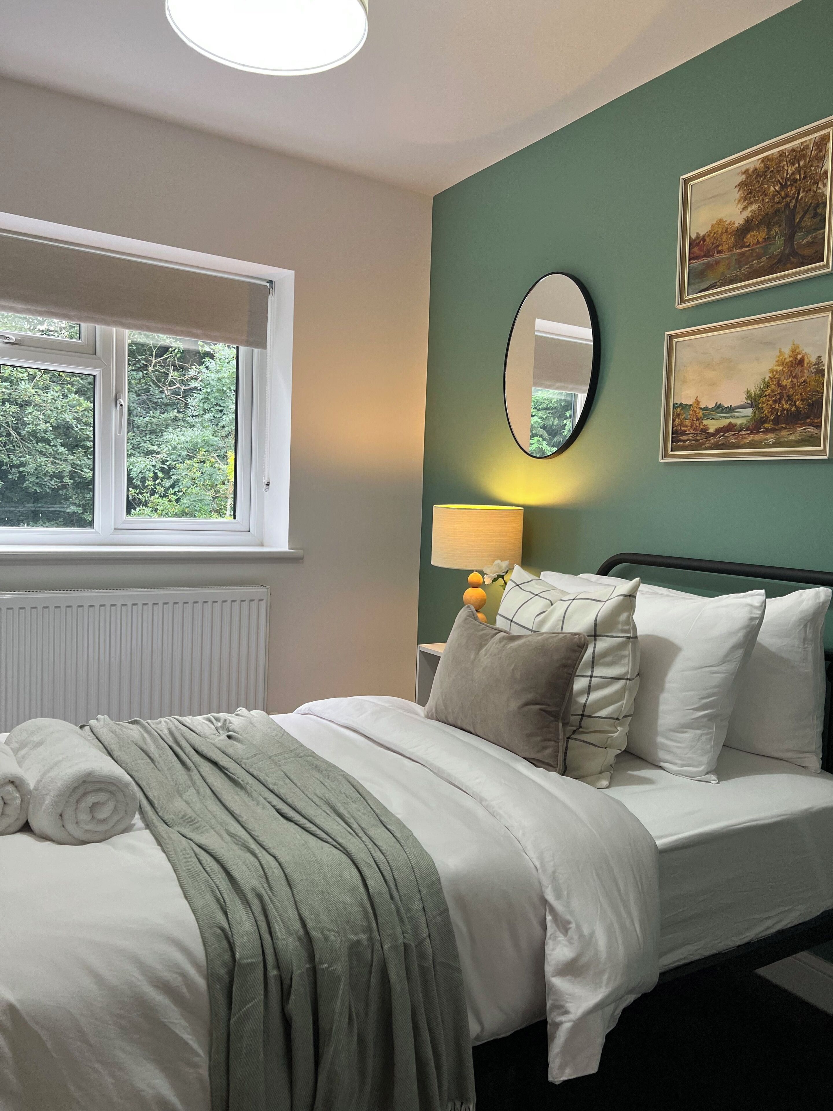 A neatly made bed with white linens and a green throw, a bedside lamp, and two framed landscape paintings on a teal wall.