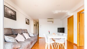 A living room with a grey sofa, white dining table, and chairs. There is a TV on the wall and a door with a wooden frame.