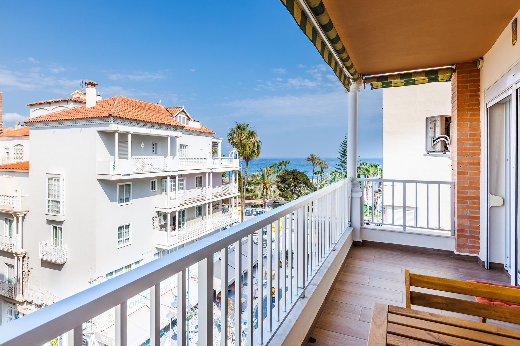 A balcony with a wooden bench, overlooking a coastal area with buildings and palm trees.